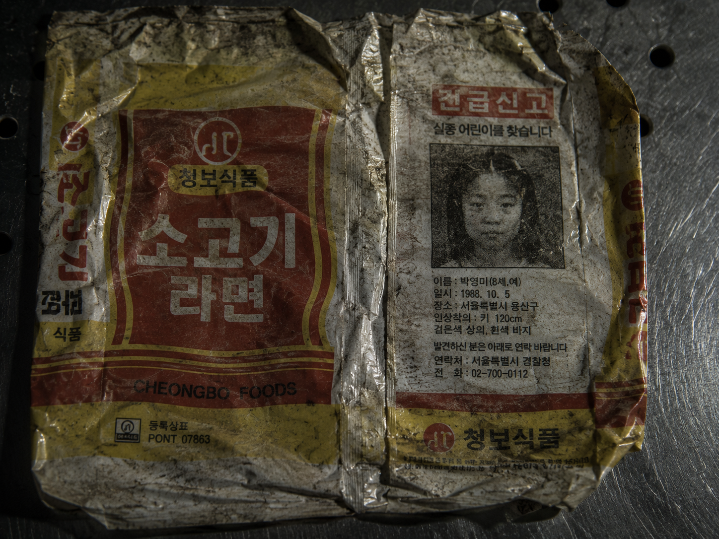 A faded, crumpled 1980s Korean Cheongbo Foods ramyun wrapper showing a grainy missing person print on a metallic background under harsh forensic lighting.