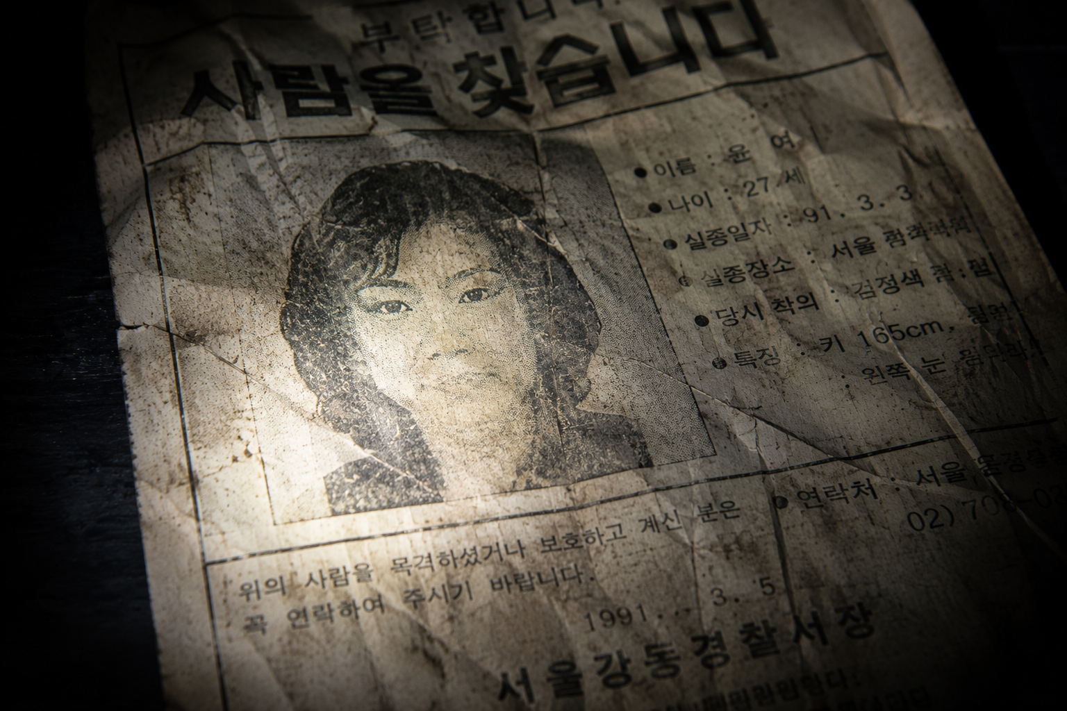Macro close-up of a damaged 1991 paper missing persons flyer illuminated by a direct flashlight beam in a dark archival room.