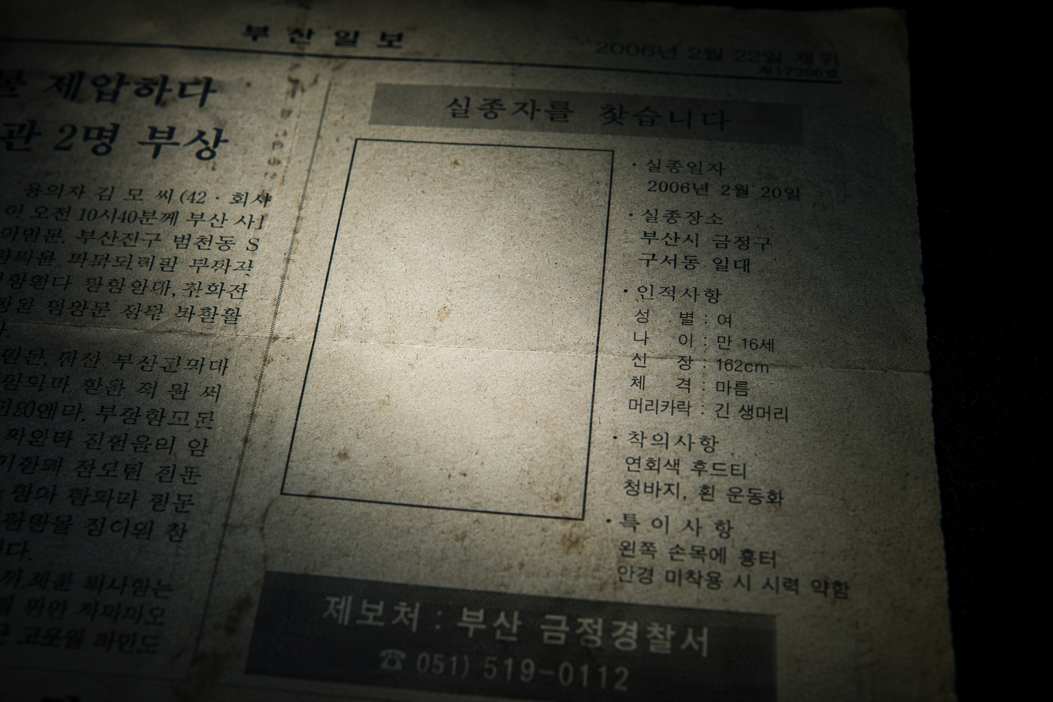 Macro close-up of a faded 2006 Korean newspaper document showing an empty missing person profile image box under a directional flashlight.