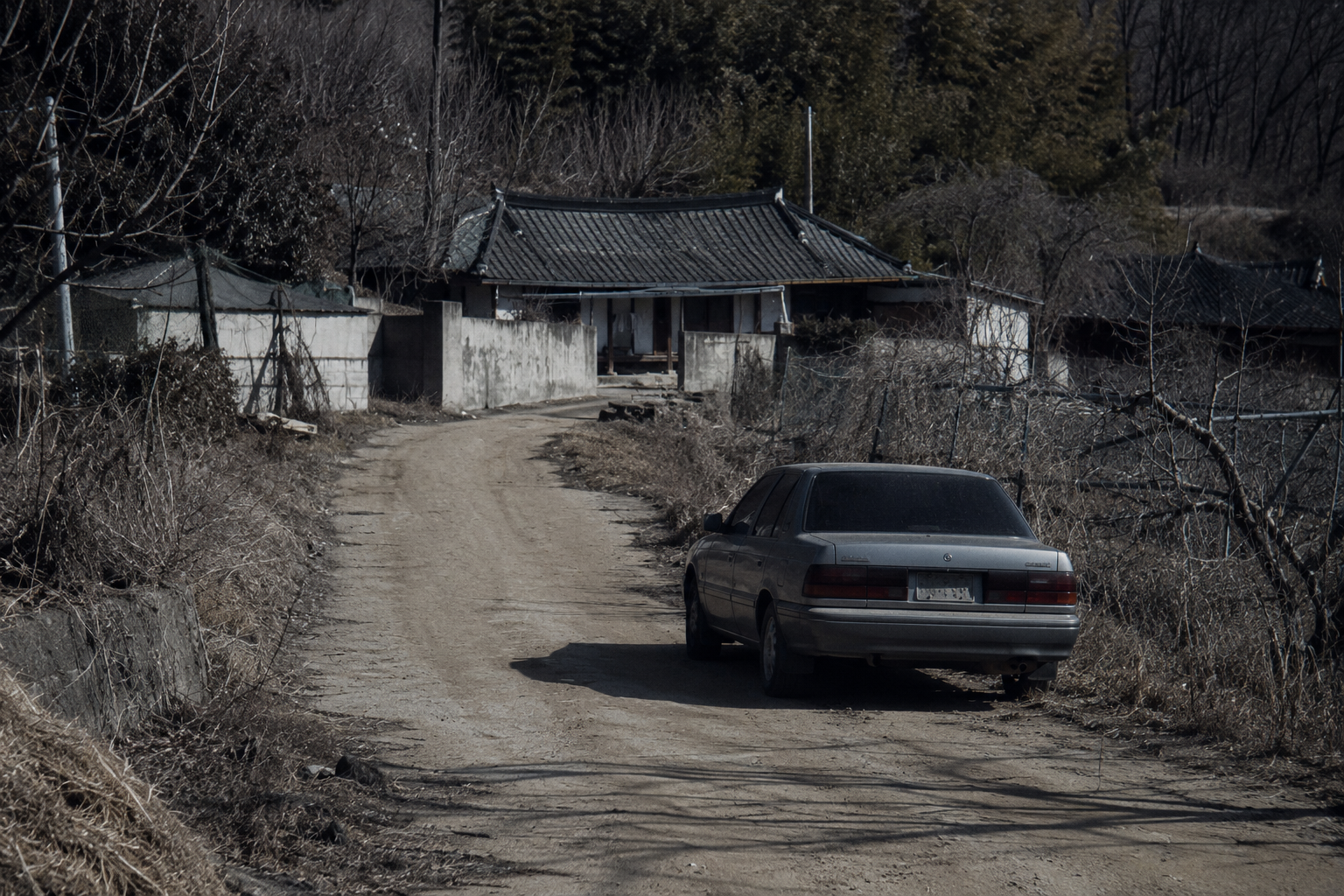 Cold forensic landscape photography of an isolated rural South Korean dirt road with a phantom 1990s mouse-grey sedan parked outside a peripheral house under bleak daylight.