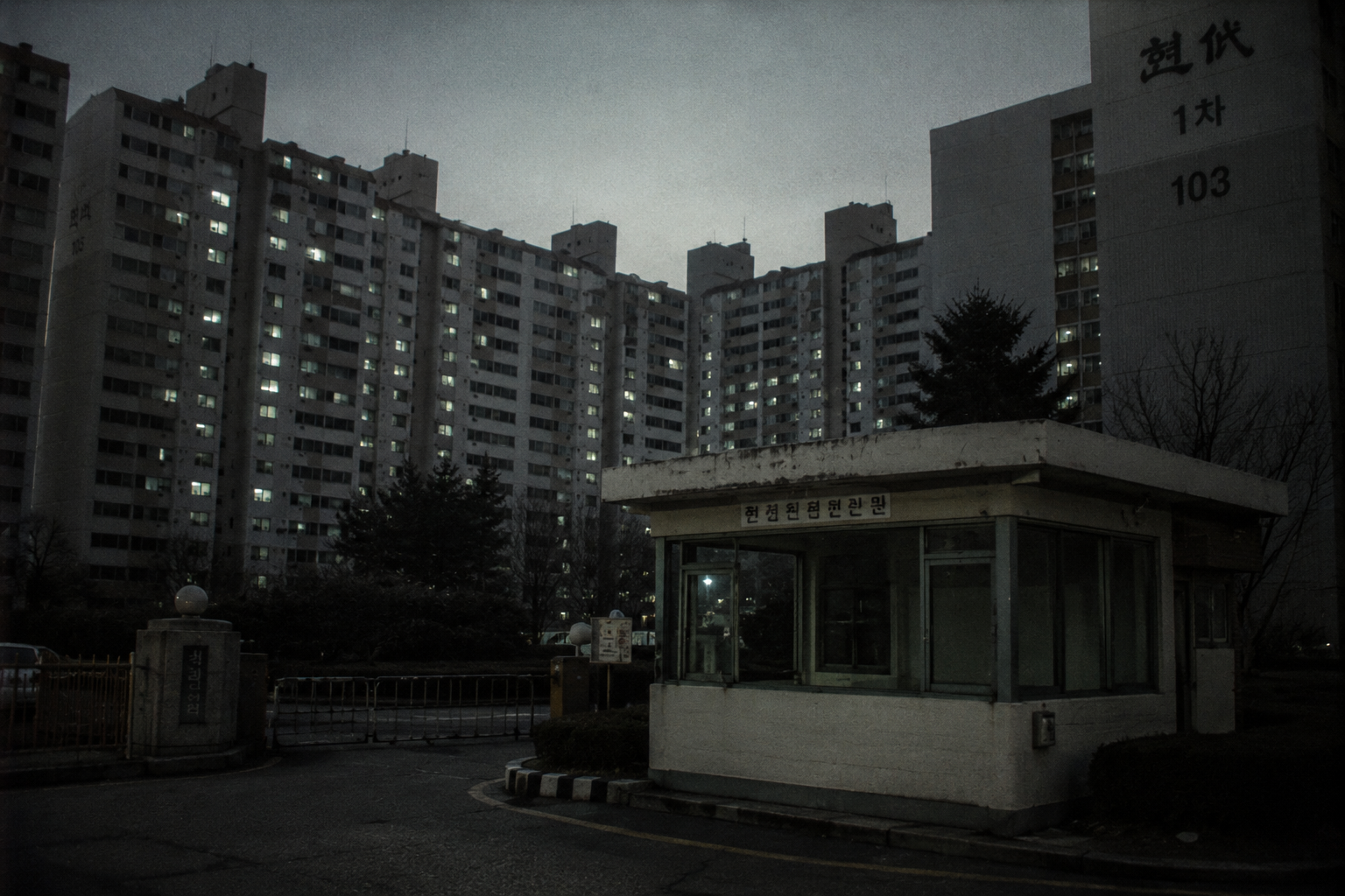 An empty concrete apartment gate and unmanned security guard booth under dim fluorescent twilight in a 1999 South Korean residential complex.