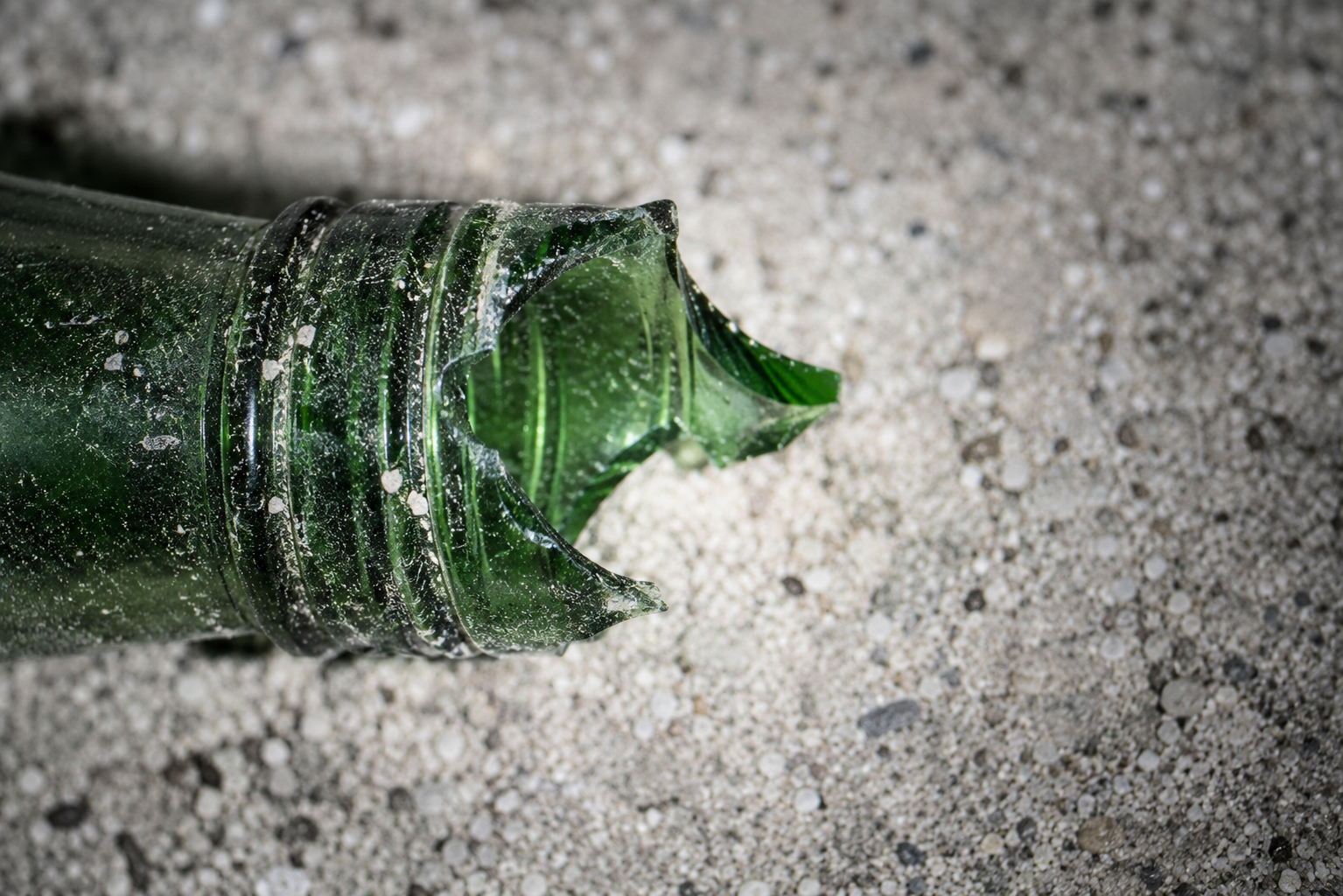 Forensic macro detail shot of a fractured green glass bottle neck on dirt ground under the direct beam of an investigative flashlight at a crime scene.