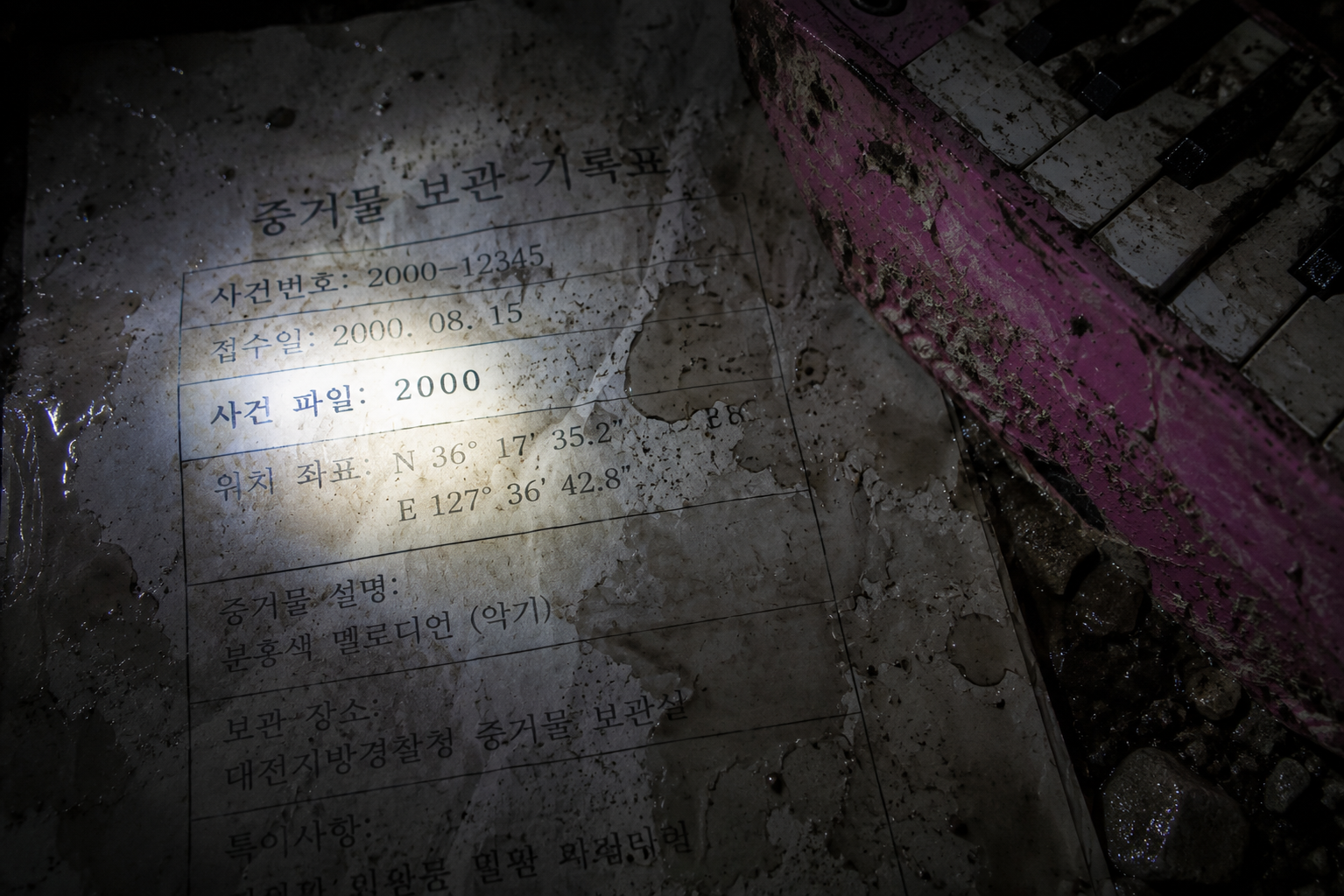 A close-up forensic capture of a water-damaged 2000 police report alongside a silt-covered pink plastic instrument under a harsh flashlight in a dark archive.