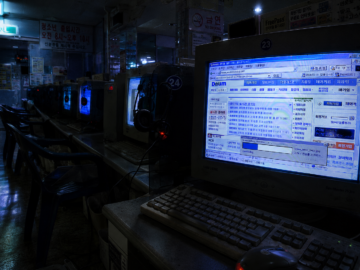The God of the Korean Darknet: The Anonymous Uploader Who Controlled 70% of a Nation’s Forbidden Media A vacant, dimly lit mid-2000s Korean internet cafe with old CRT monitors glowing in a dark room, capturing an eerie early net archive atmosphere.