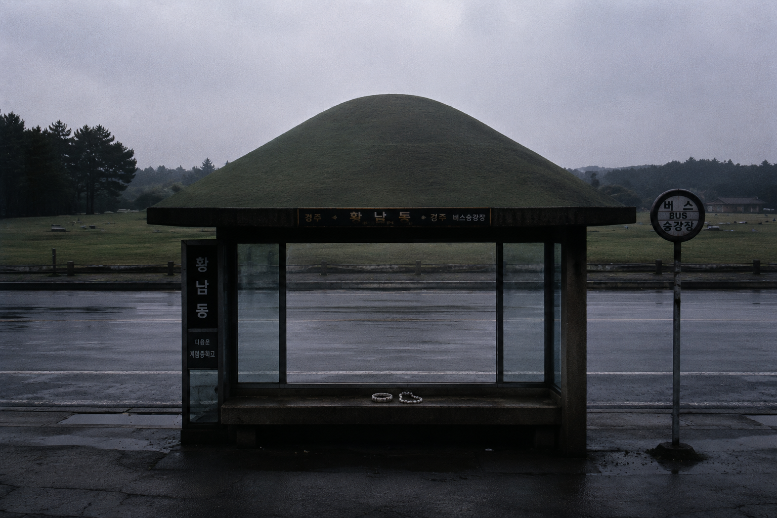 An empty 1991 Gyeongju bus stop facing a massive ancient grassy burial mound under a misty sky, with an abandoned jade ring on the bench.