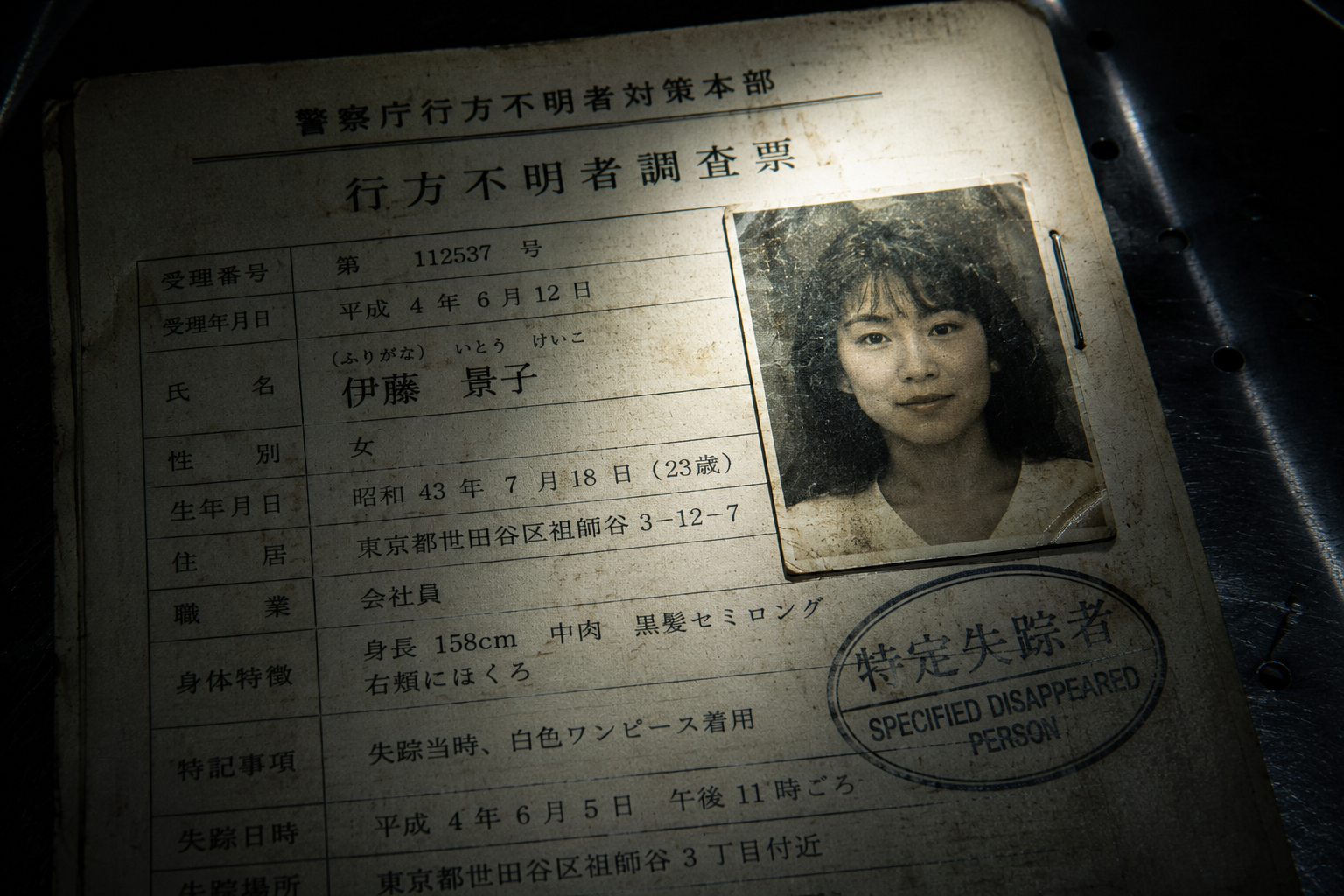 A macro close-up of a faded 1990s Japanese police file for a missing person, illuminated by a flashlight on a dark metal desk.